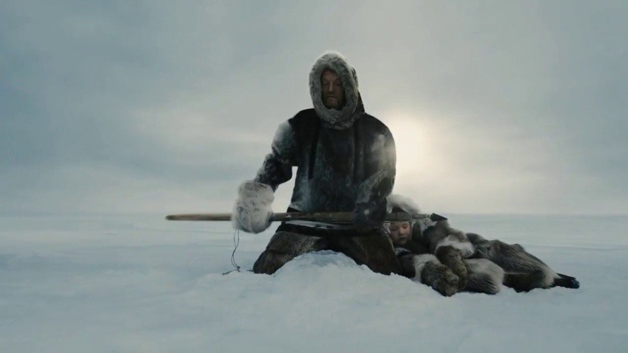 A man wrapped in furs sits by a hole in the ice holding a spear, as a child also wrapped in furs sleeps with their head on his lap