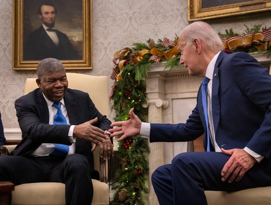 Two smiling men wearing suits reach out to shake hands while seated.