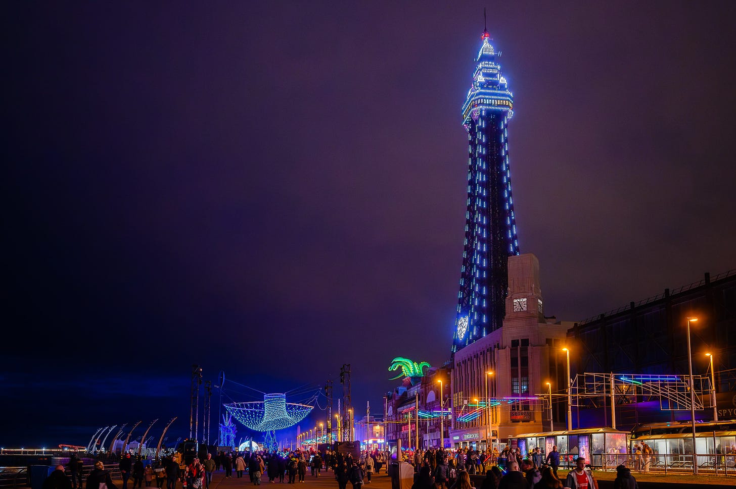 Blackpool Promenade at dusk. A net of colourful LED lights hangs over the prom as people walk beneath. In the distance Blackpool Tower is lit up.