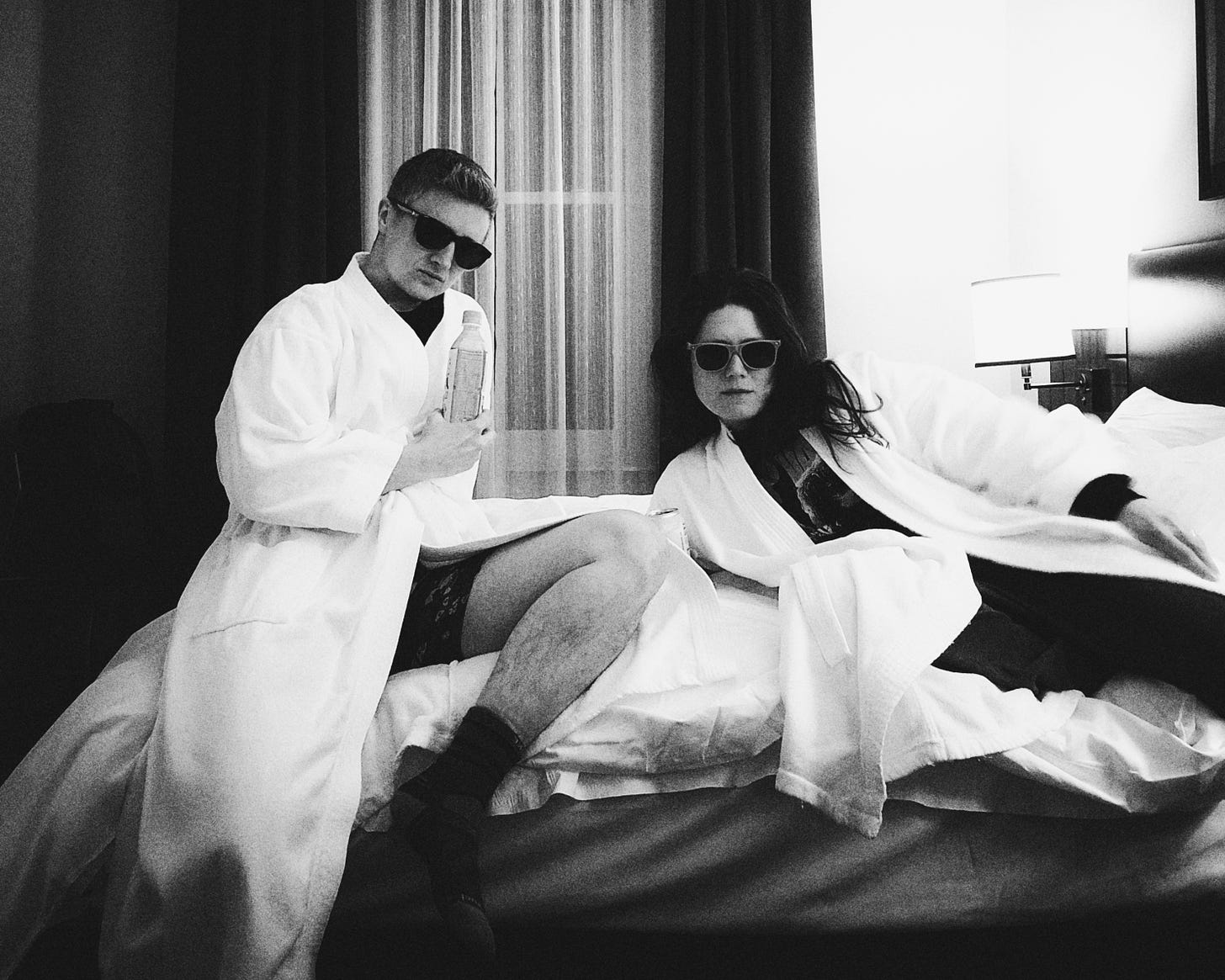 Black and white photo of Emily and Meg, both wearing sunglasses and serious faces, lounging on a hotel bed while dressed in fluffy white robes.