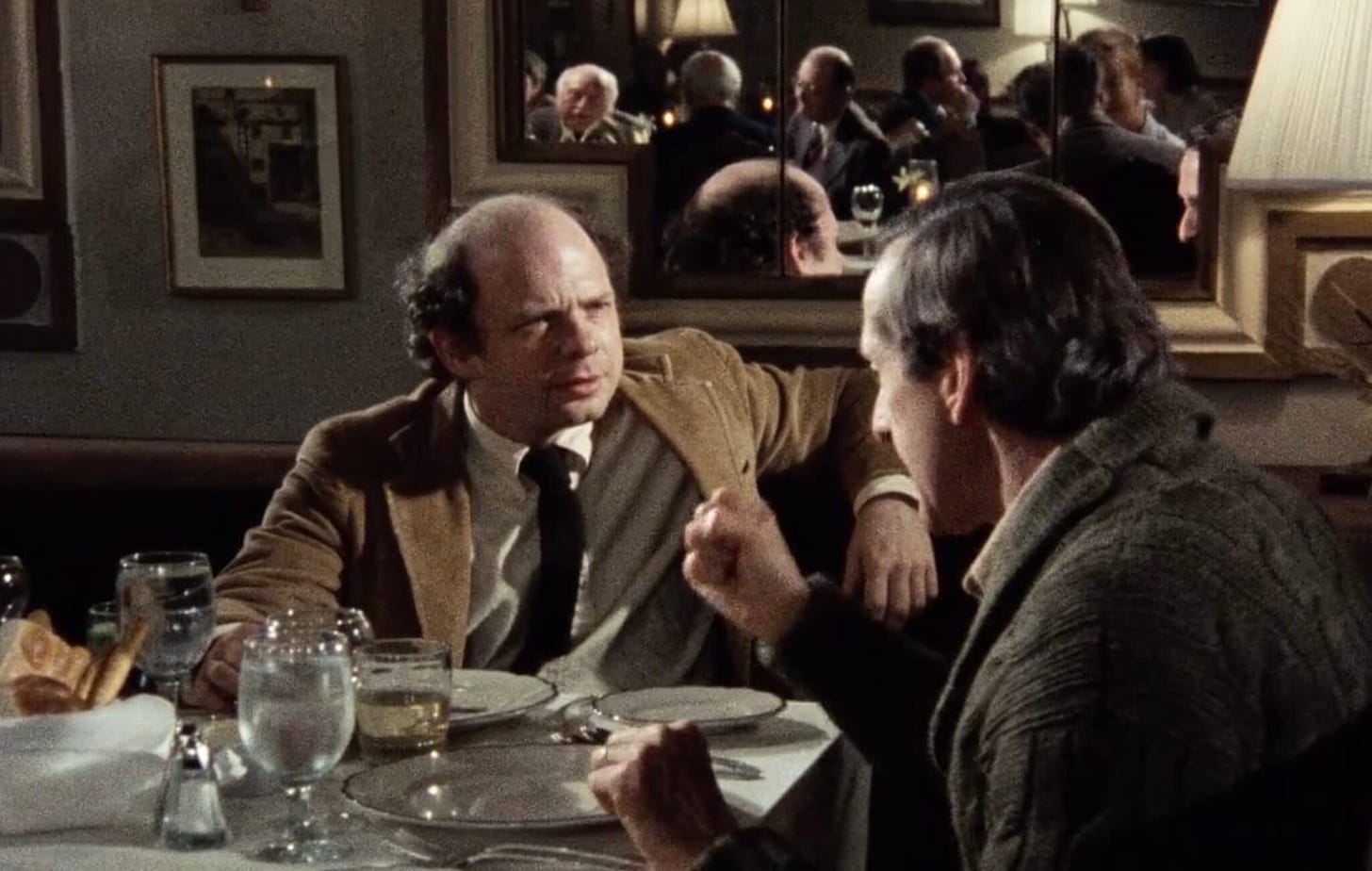 Screencap from My Dinner With Andre. Wallace Shawn listens, his arm resting on the back of the chair. Andre is telling a story. No food on their plates.