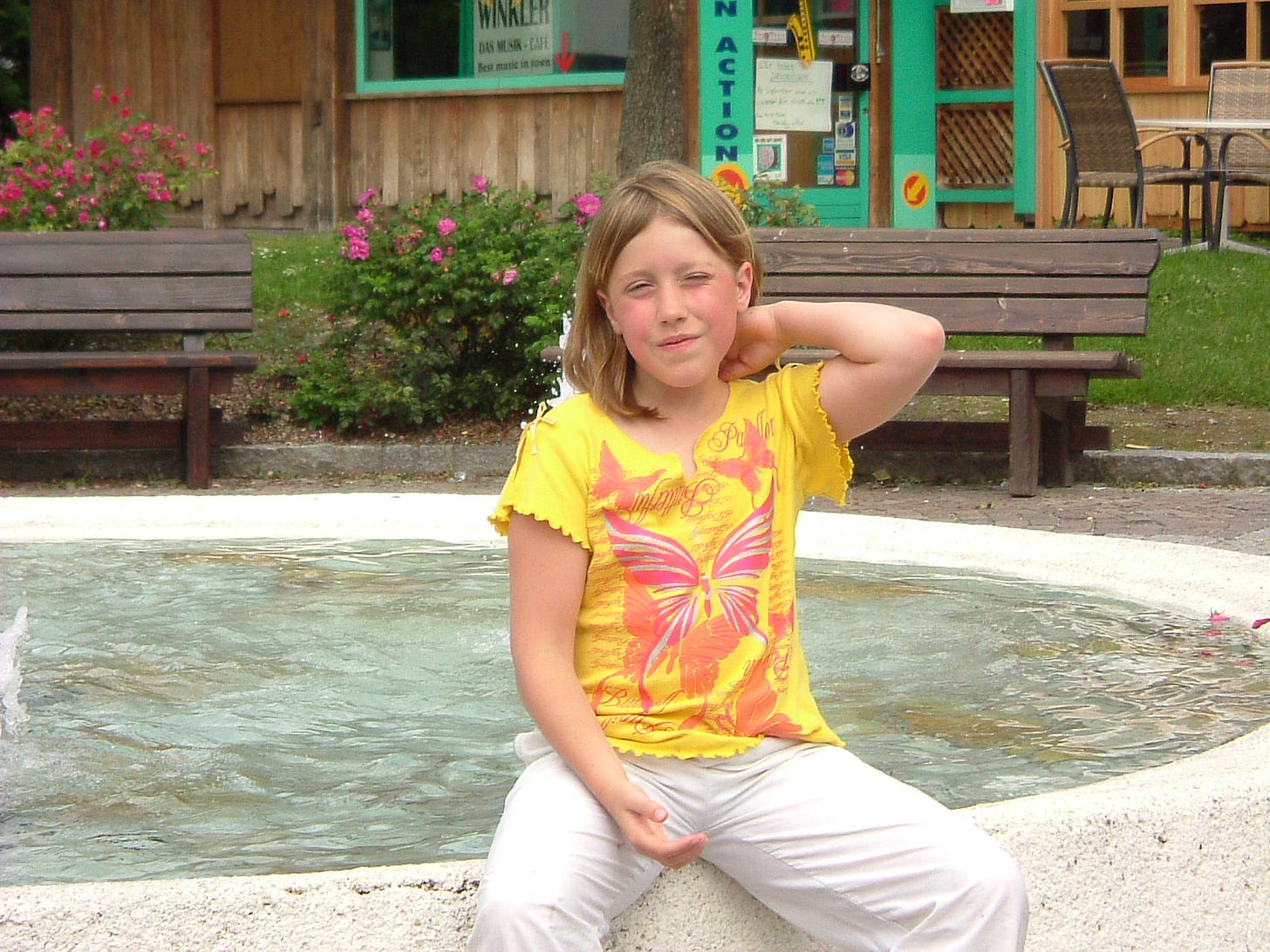 A girl with a yellow t-shirt with butterflies sitting on the edge of a fountain while squinting her eyes, touching her neck, with a red, tired face