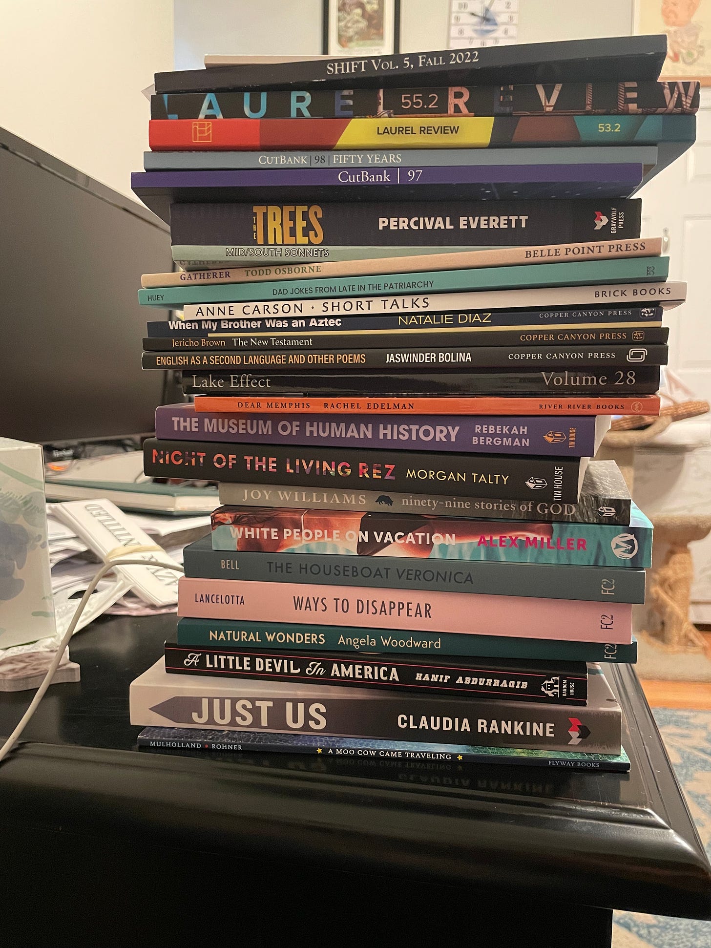 a stack of books, too towering to name them all, sitting on a desk with a cat tower visible in the background