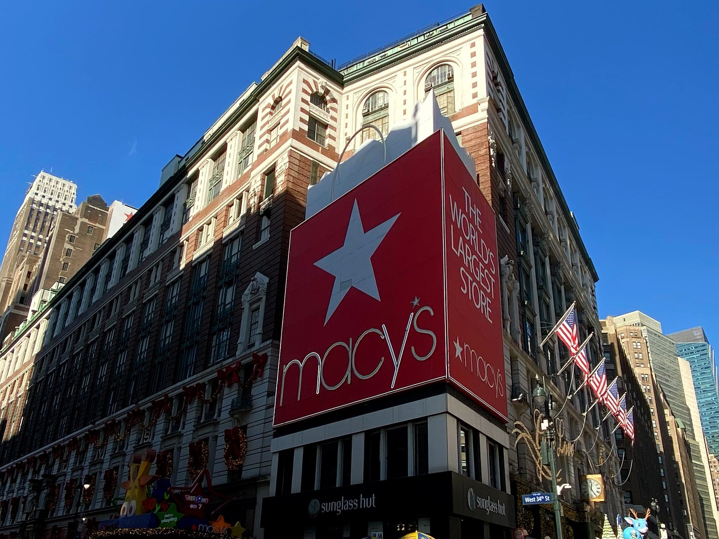 The corner of 34th and Broadway. The Macy's building is clearly designed around a carved out corner lot, currently housing a Sunglasses Hut. There is a large billboard in the shape of a Macy's shopping bag above it.