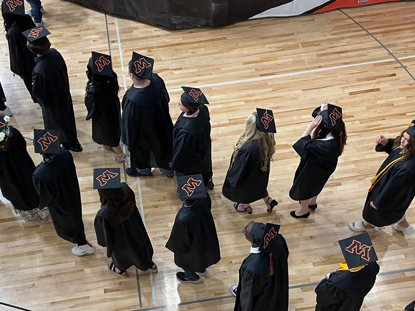 Young people in caps and gowns process across a hardwood floor
