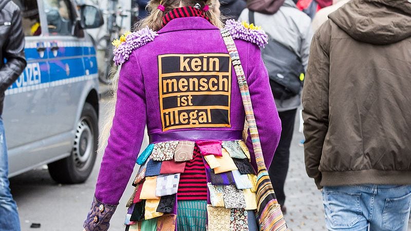 The back of a protestor in a bright purple jacket with a patch that reads "kein mensch ist illegal" meaning no person is illegal.