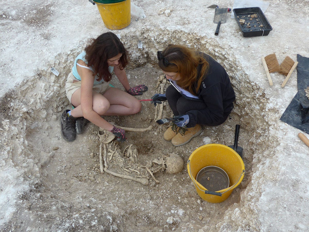Two women in a cricular burial pit containing a specimen, surrounded by archaeological tools