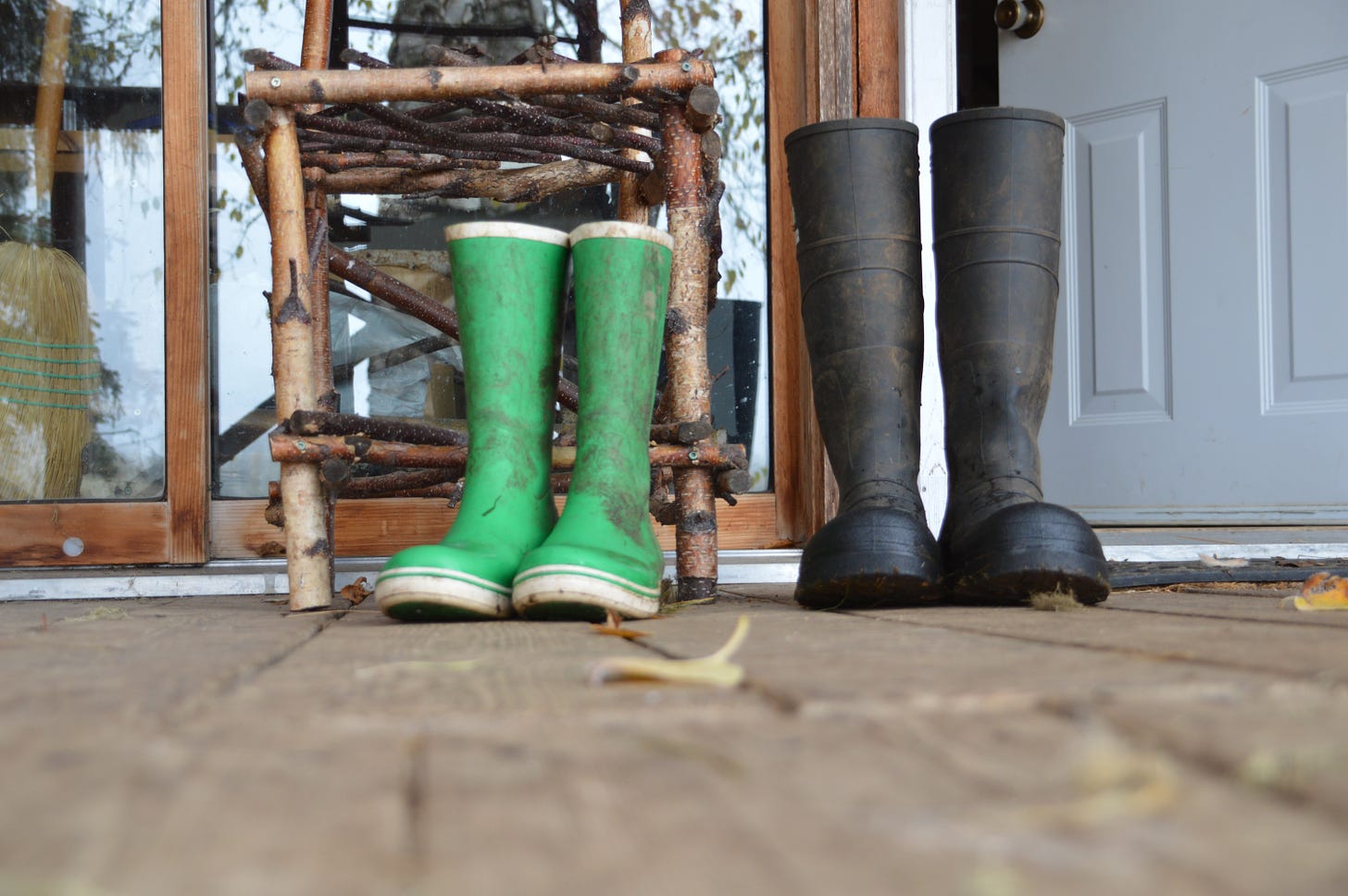 Two pairs of rubber boots - green and black - stand in front of a wooden chair, outside a doorway entrance