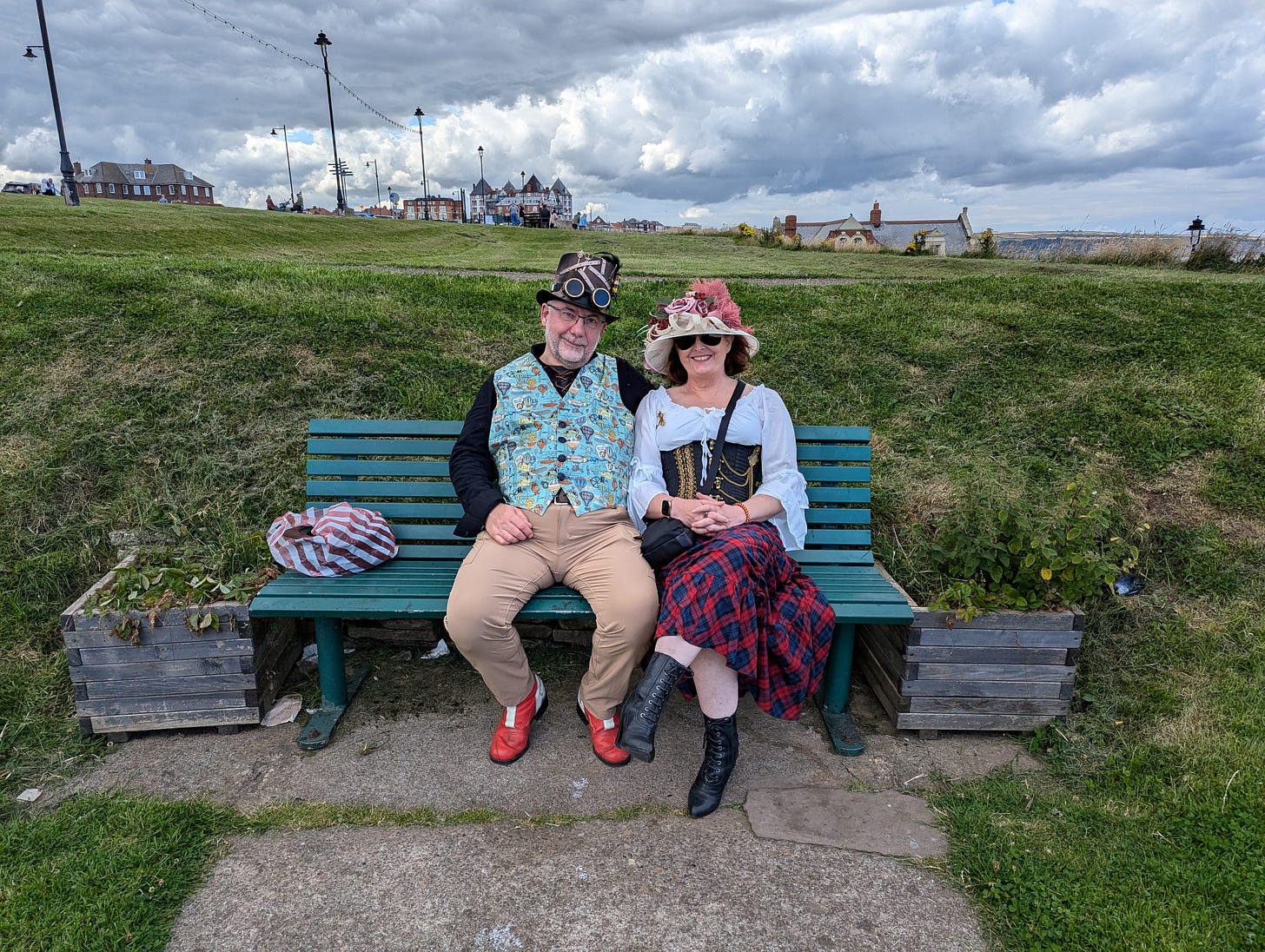 A steampunk couple sit on a bench, smiling into the camera