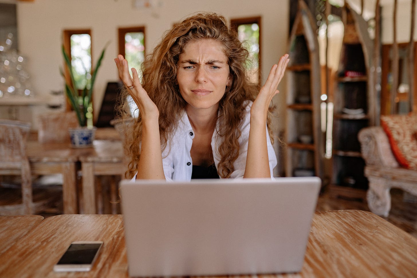 A woman with long, wavy hair sits at a desk with her laptop. Her hands are up and she has a look of confusion on her face. 