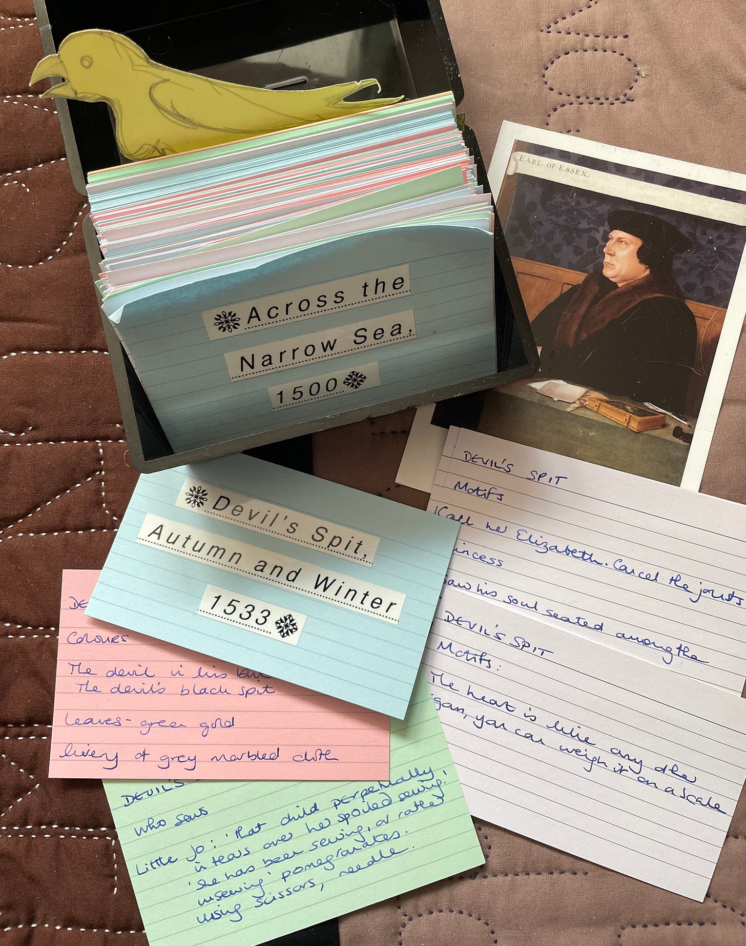A box of index cards, open at a card that reads “Across the Narrow Sea, 1500”; a yellow bird cut out of card; a postcard of Thomas Cromwell. Some index cards are scattered outside the box.