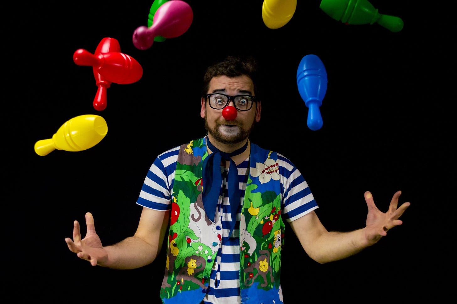 stock image of a white guy in glasses and wacky clown outfit failing to juggle multi-colored plastic bowling pins