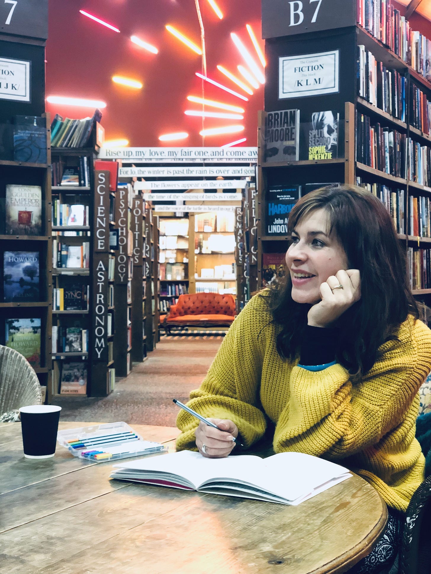 Claire, a white woman with dark hair sits in at a table in a second hand book shop with a notebook and pen.  She is smiling and looking away from the camera. There are red and orange and firework inspired lights behind her.   