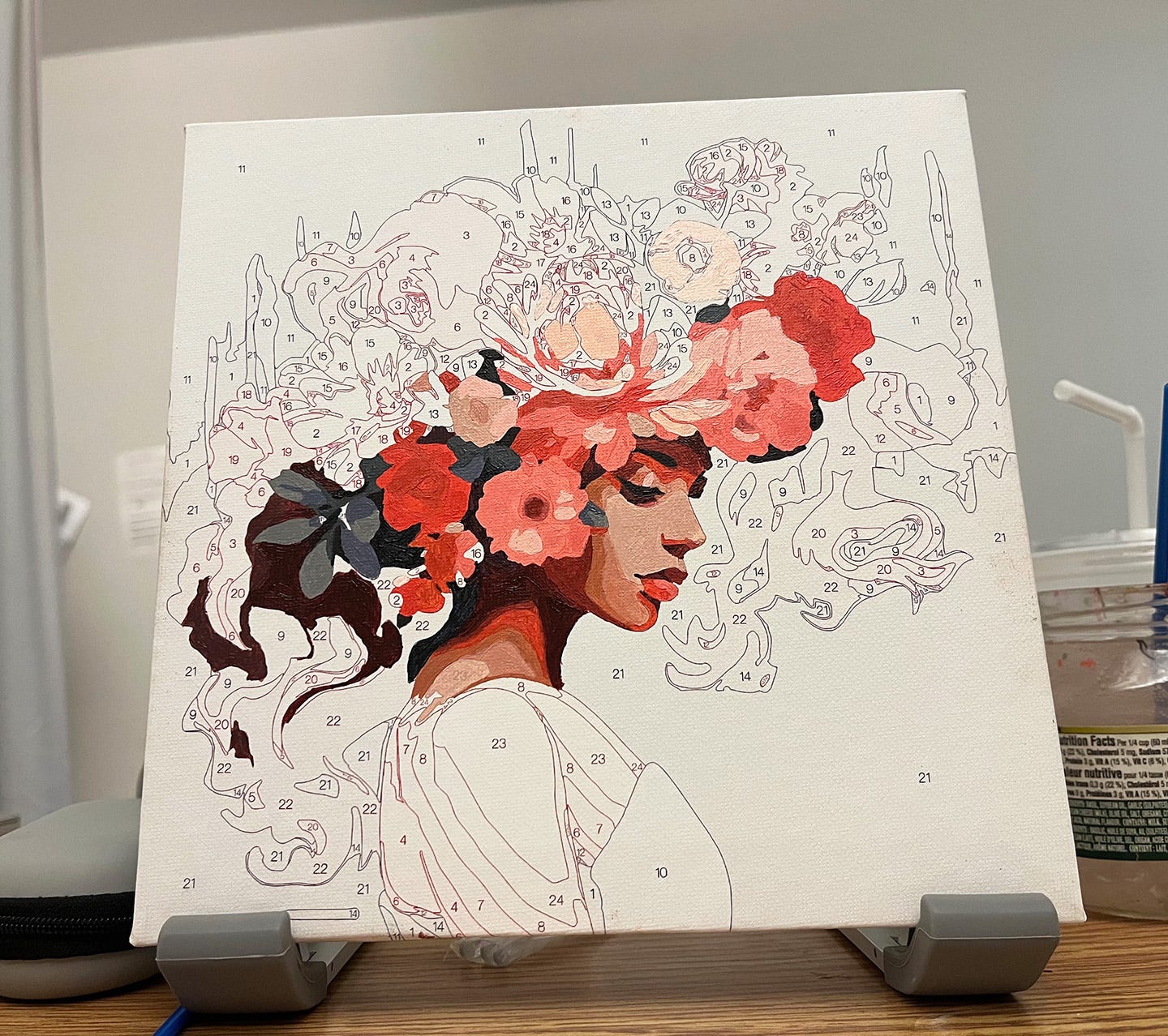 Partially completed paint-by-number propped up on a hospital tray table. The painting shows a woman in profile with enormous mass of large pink and red flowers woven into her hair.