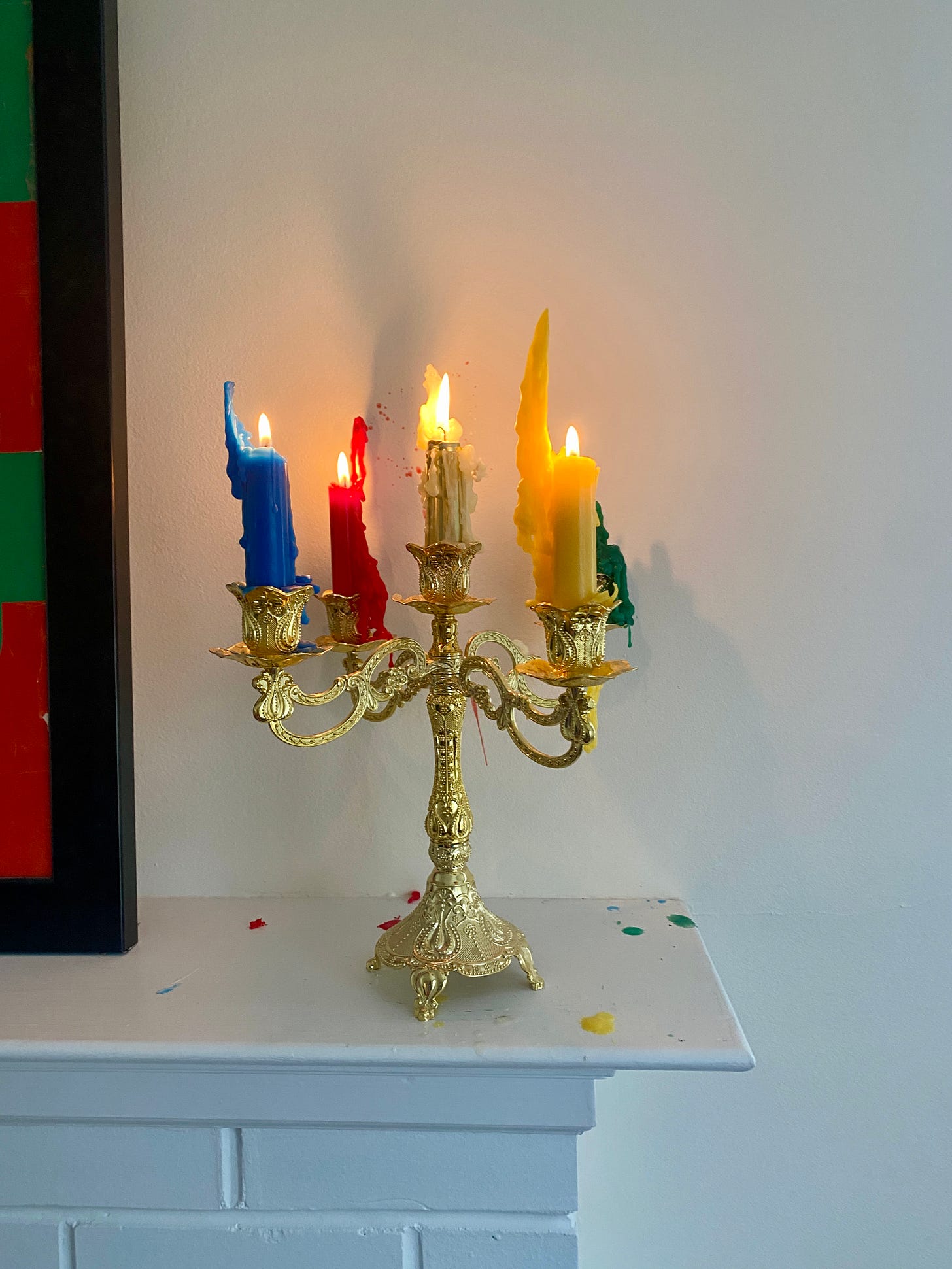 A photo of a gold candalabra sitting on a white fireplace mantle. In the candle holder there are five candles — red, blue, green, yellow, and gold. They are all lit taper candles that have burned 2/3rds of the way down. There are wax drops and drippings on the wall and the mantle.