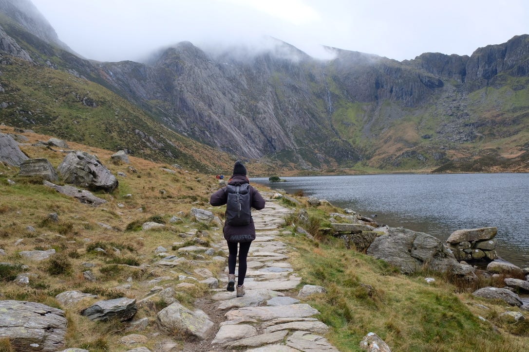 A woman in a black hat and black coat and leggings walks on a rock path toward a mountain, with a lake to her right.
