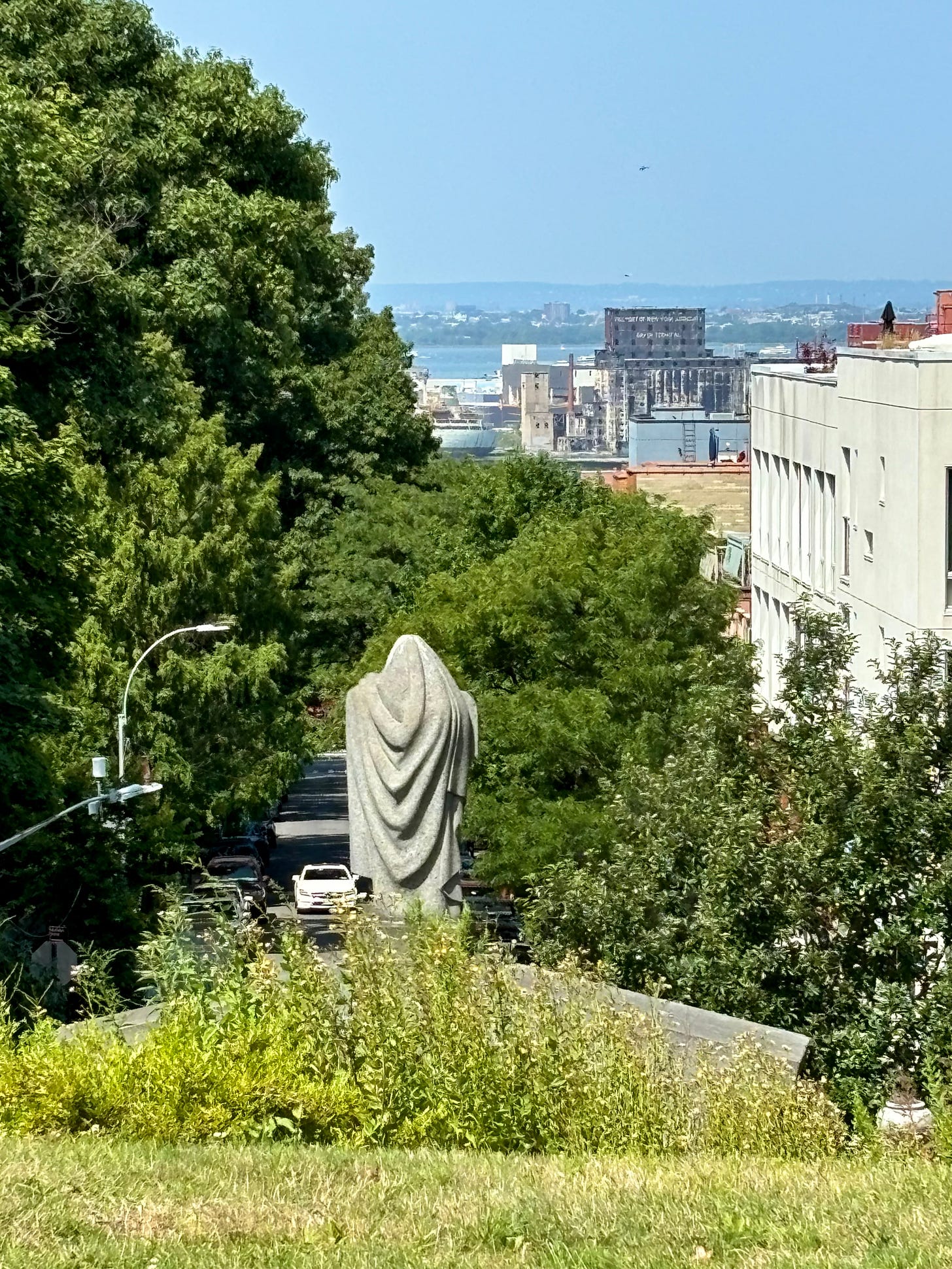 Looking down a hill towards Brooklyn. There is a cloaked statue facing the city. In the distance are warehouses.