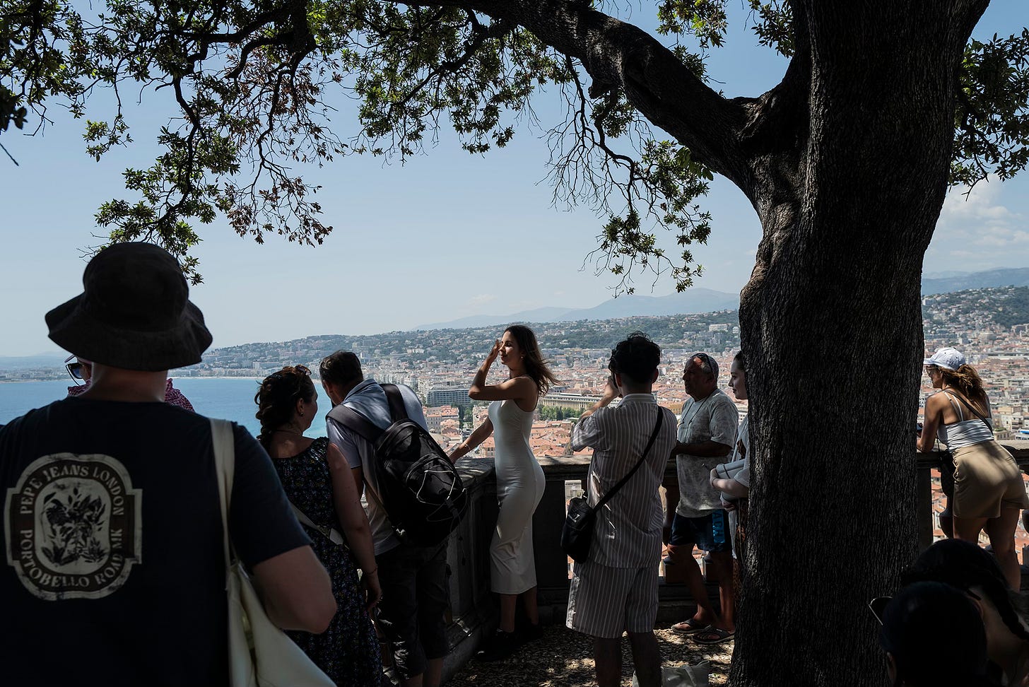 A group of people gathered under a tree, gazing at the scenic view of Nice, France, from an elevated vantage point. Shadows and silhouettes contrast against the bright coastal landscape.