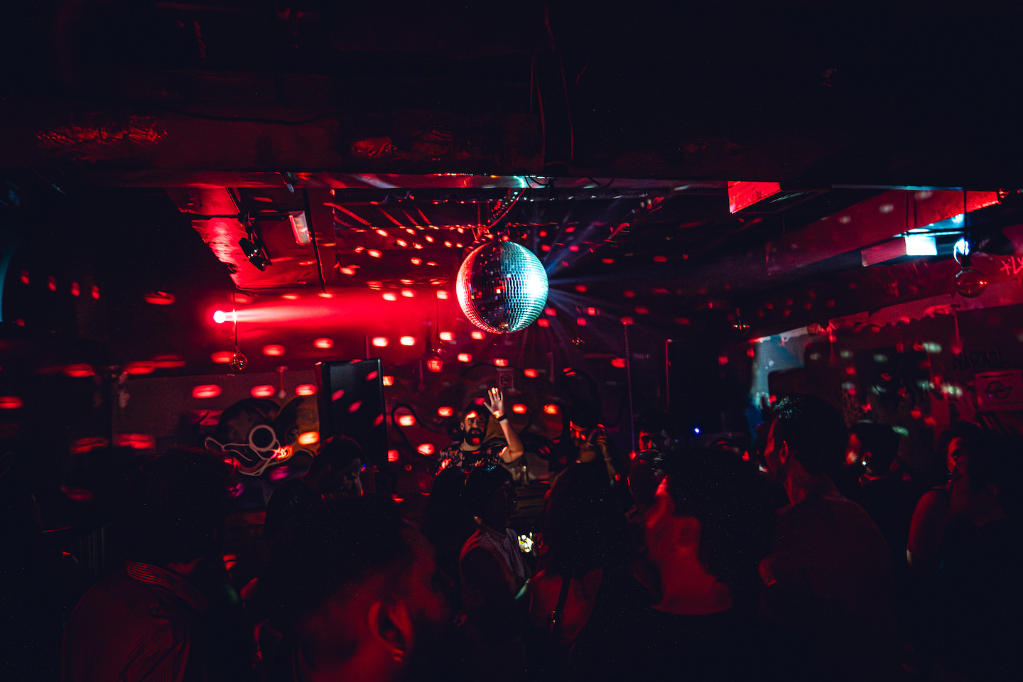 Imagem: Pista de festa escura, com luz vermelha e um globo de espelhos no meio. Na frente se vê um DJ levantando a mão para o público.