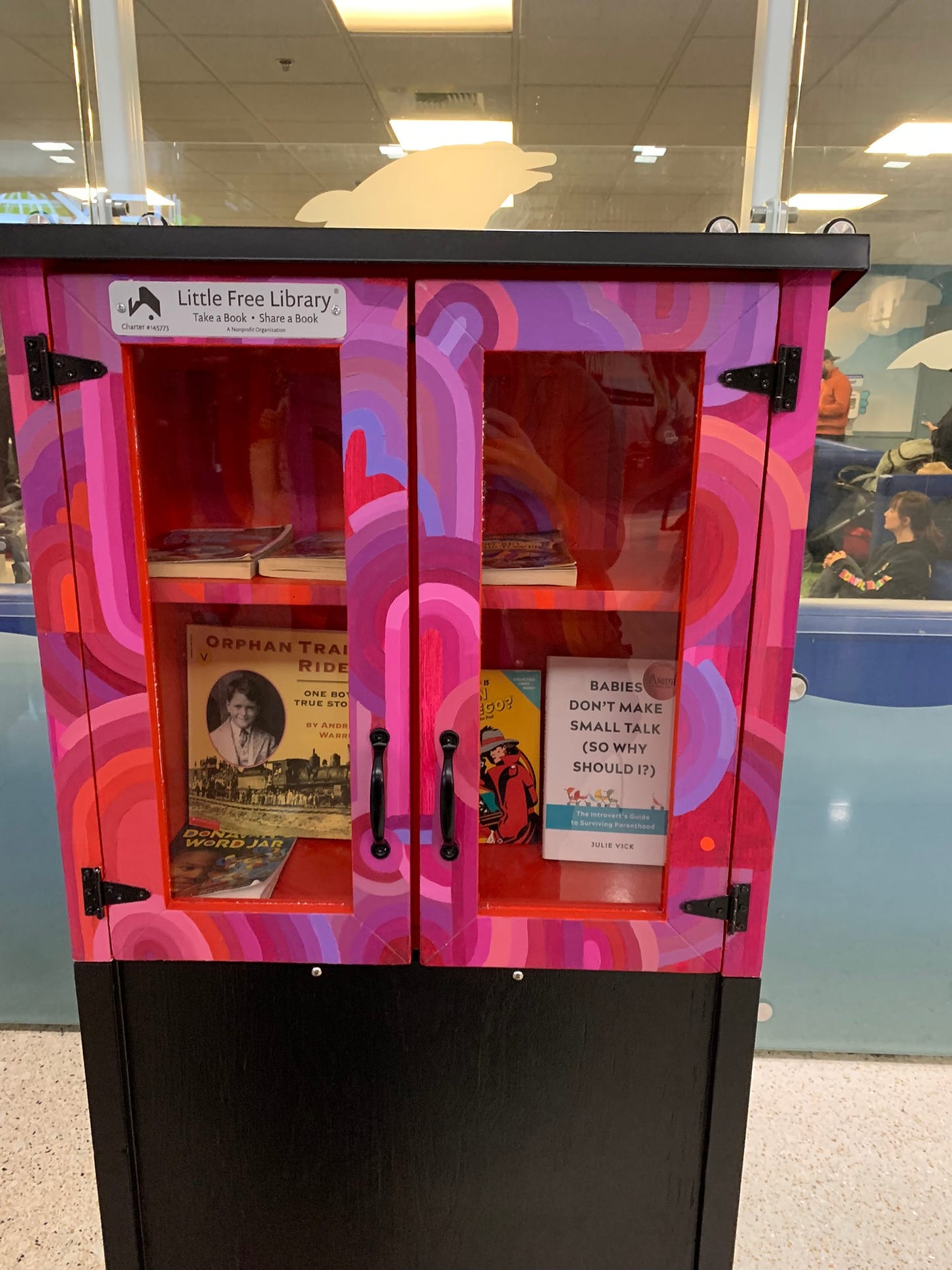 A photo of my book in an artistically decorated little free library at the Seattle Airport