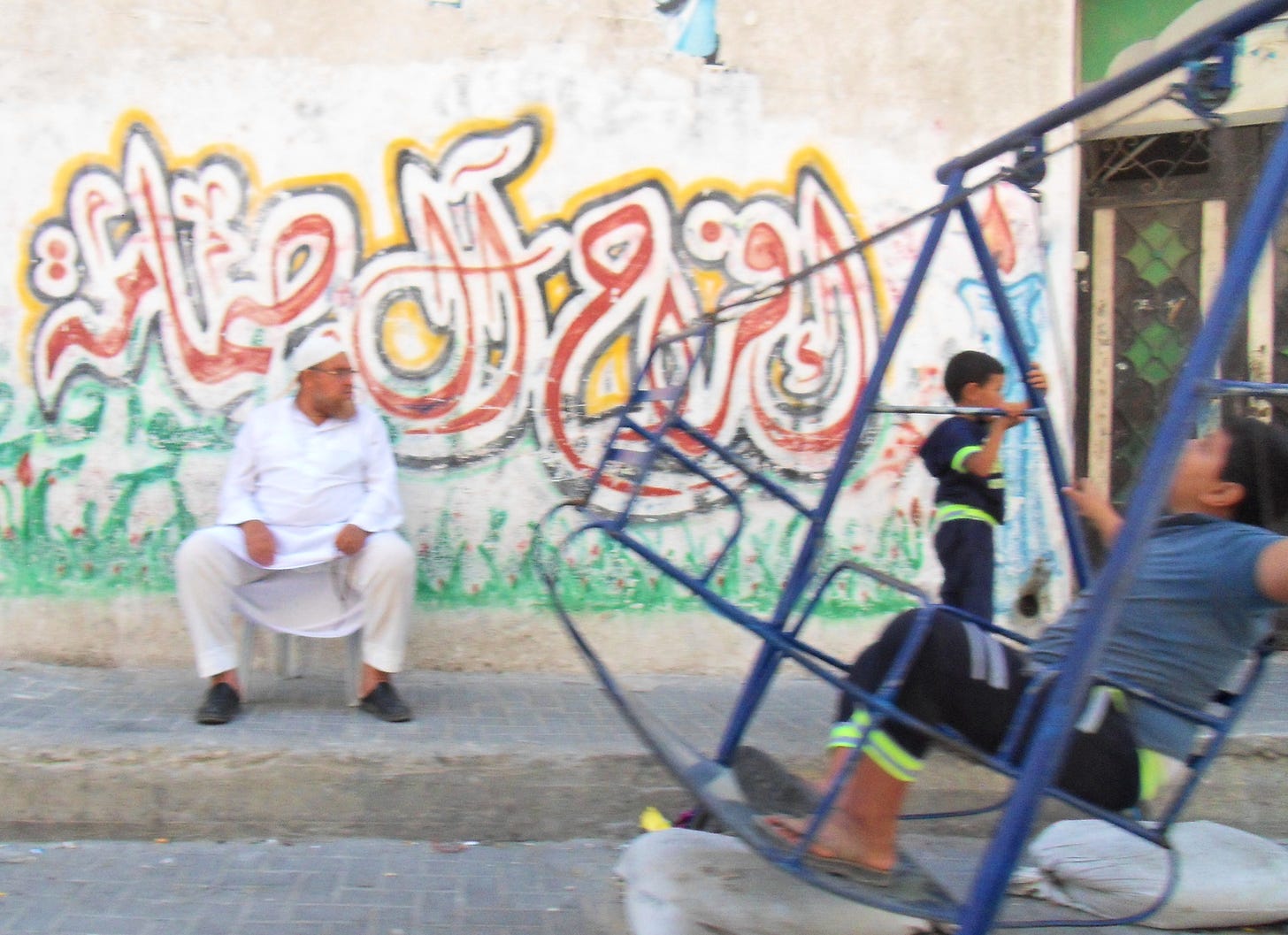 A man watches as two boys play on a blue swingset in front of a graffitied concrete wall