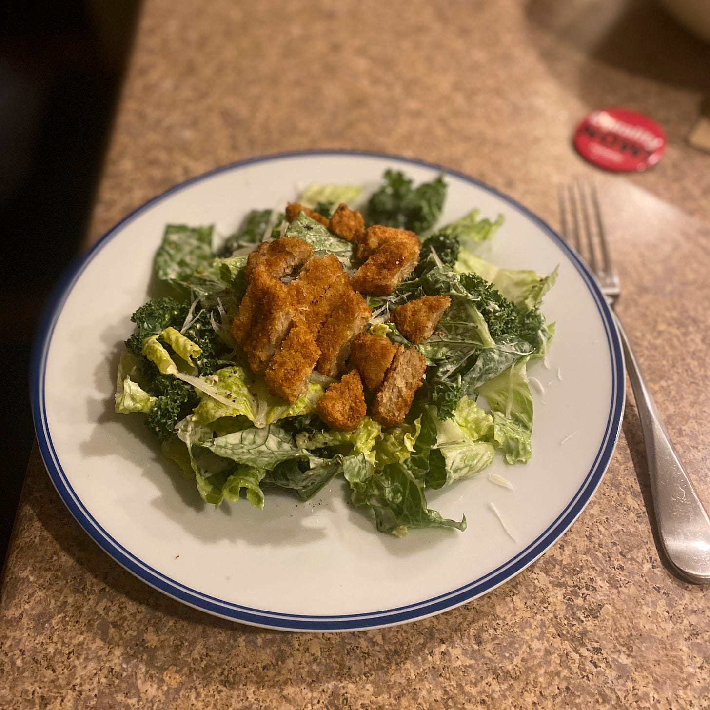 A white blue-rimmed plate with the kale and romaine caesar described above, with sliced vegan chick'n tenders on top. A fork is beside the plate and there is a blurred badge in the background that reads 'equality now'