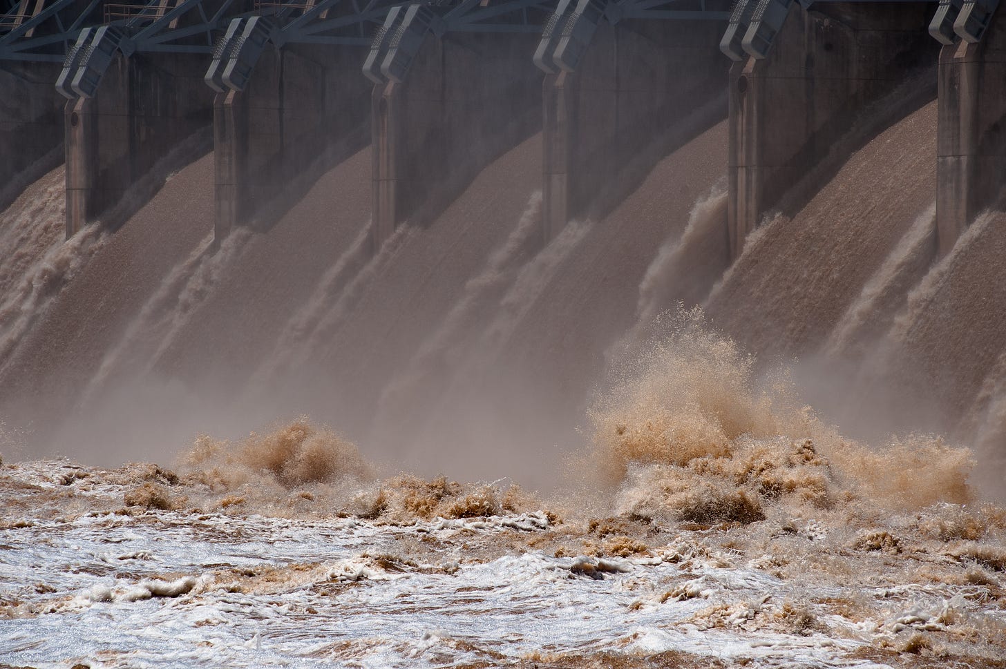 Seen from just downstream, the frothing, muddy waters of Keystone Reservoir roar through the gates of Keystone Dam between massive concrete piers. The water crashes into the churning Arkansas River in a furious explosion.