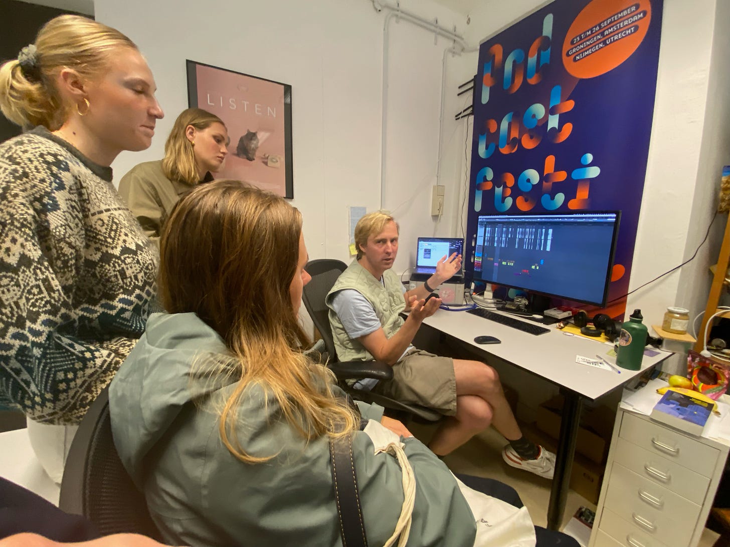 Foto van Lieven aan zijn bureau op zijn kantoor, hij heeft blond haar en een groene bodywarmer aan. Hij zit voor een groot scherm waar een montage op te zien is. Drie blonde meisjes kijken gefascineerd naar hem. Op de muur een banner van het Podcastfestival 2021 en een poster van de film LISTEN.