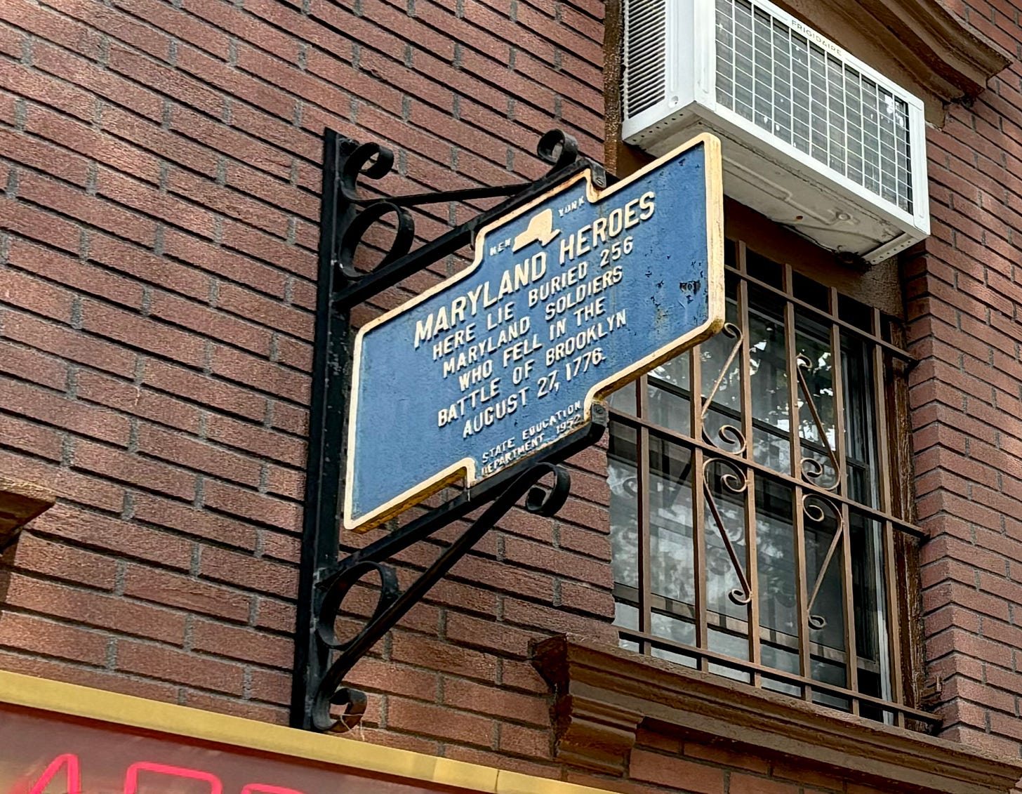 A navy sign with cream writing hanging from the American Legion. It reads: Maryland Heroes, Here lie buried 256 Maryland Soldiers who fell in the Battle of Brooklyn, Aug. 27, 1776.