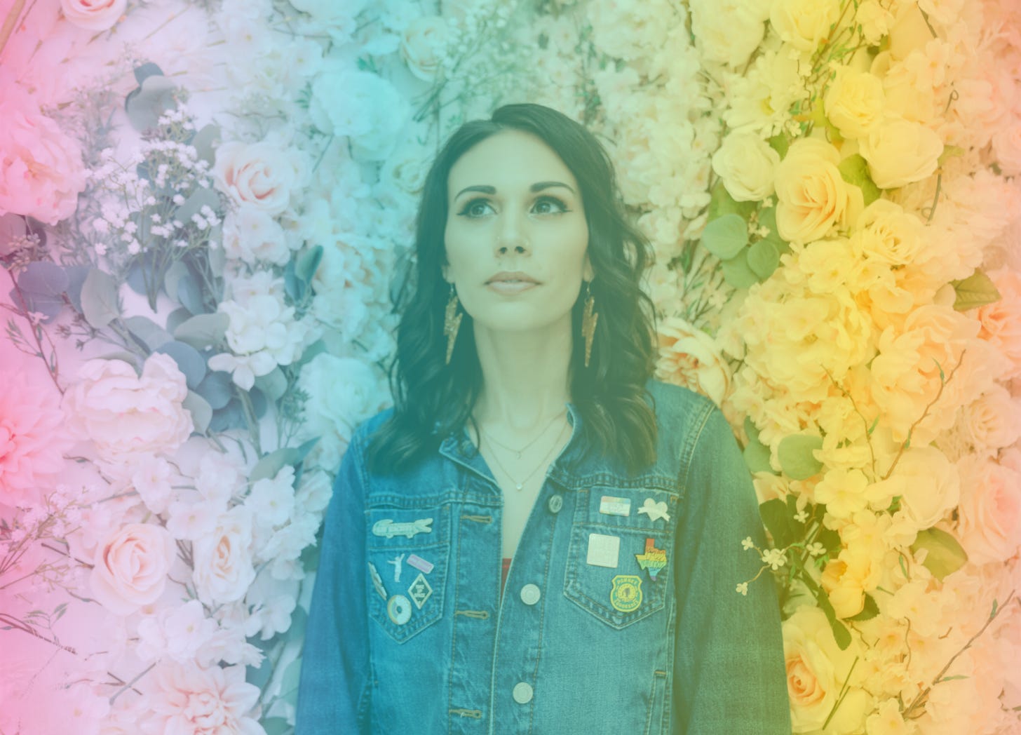 A rainbow-filtered photo of Shohreh standing against a flower all of light pink flowers and greenery. She's wearing a denim jacket with enamel pins on the pockets and gold glittery lightning bolt earrings.