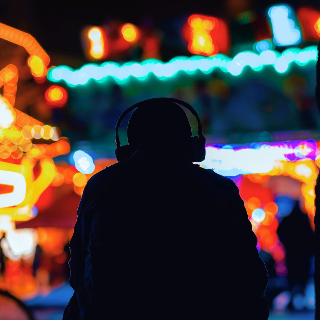 The outline of a person with old-school headphones on squatting and looking out over a multi-colored neon lights of an amusement park