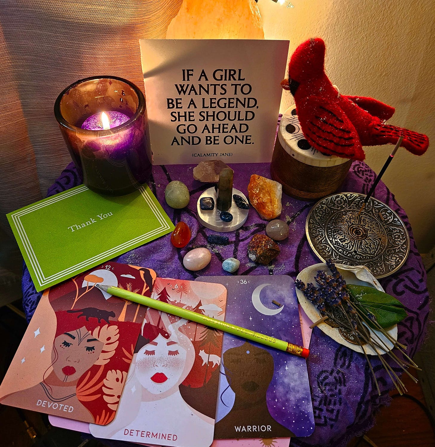 Writer Altar with candles, incense, oracle cards that read Devoted, Determined, and Warrior. Goals written in a "thank you" card, crystals, flowers, leaves, shells, and a felt cardinal. A quote by Calamity Jane reads "If a girl wants to be a legend, she should go ahead and be one."