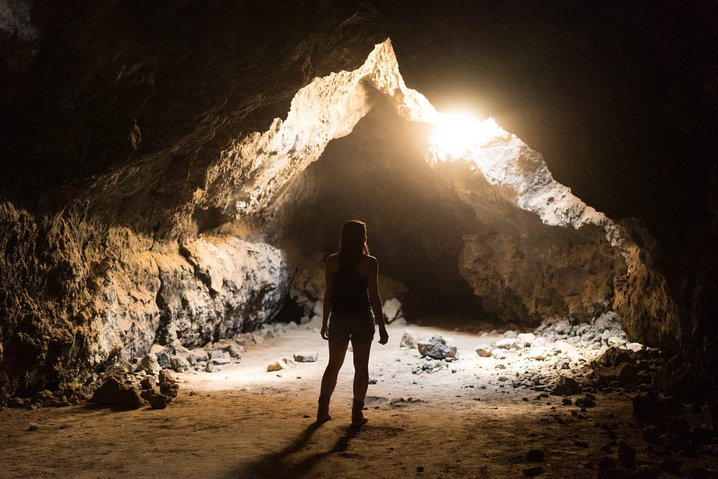 ssa imagem mostra uma pessoa de pé dentro de uma caverna, de costas para a câmera. A pessoa está olhando em direção à entrada da caverna, que é parcialmente iluminada pela luz do sol entrando do lado de fora. As paredes da caverna são rochosas e irregulares, com um tom de pedra natural, e há várias pedras espalhadas pelo chão. A iluminação cria um contraste marcante entre as partes claras próximas à entrada e as sombras mais escuras no interior da caverna. Essa composição dá uma sensação de mistério e tranquilidade.