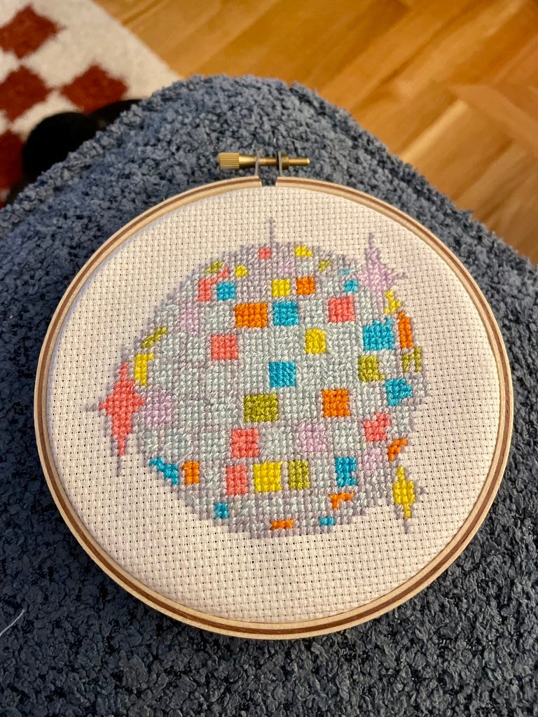 cross stich in a round hoop of a colorful disco ball on top of a blue couch with a burnt orange and white checkered rug in the background