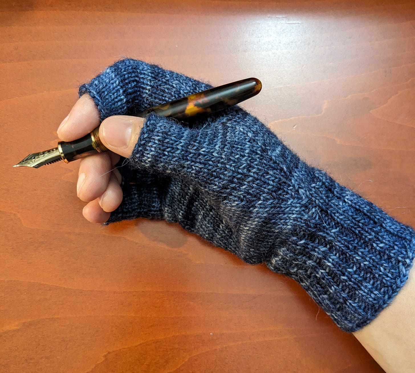 A blue hand-knit fingertip glove on a light-skinned hand holding a brown and gold fountain pen.