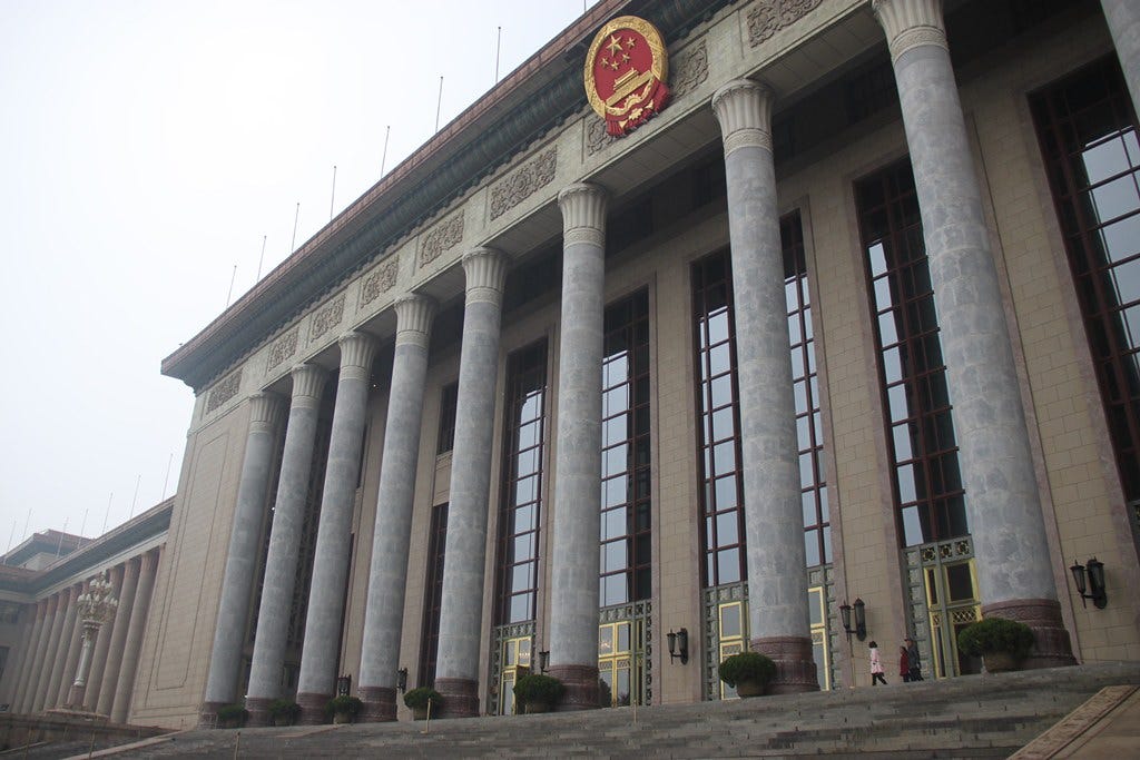 Foto colorida do Grande Palácio do Povo. Pilastras em mármore com o símbolo do partido comunista chinês no topo em vermelho e dourado