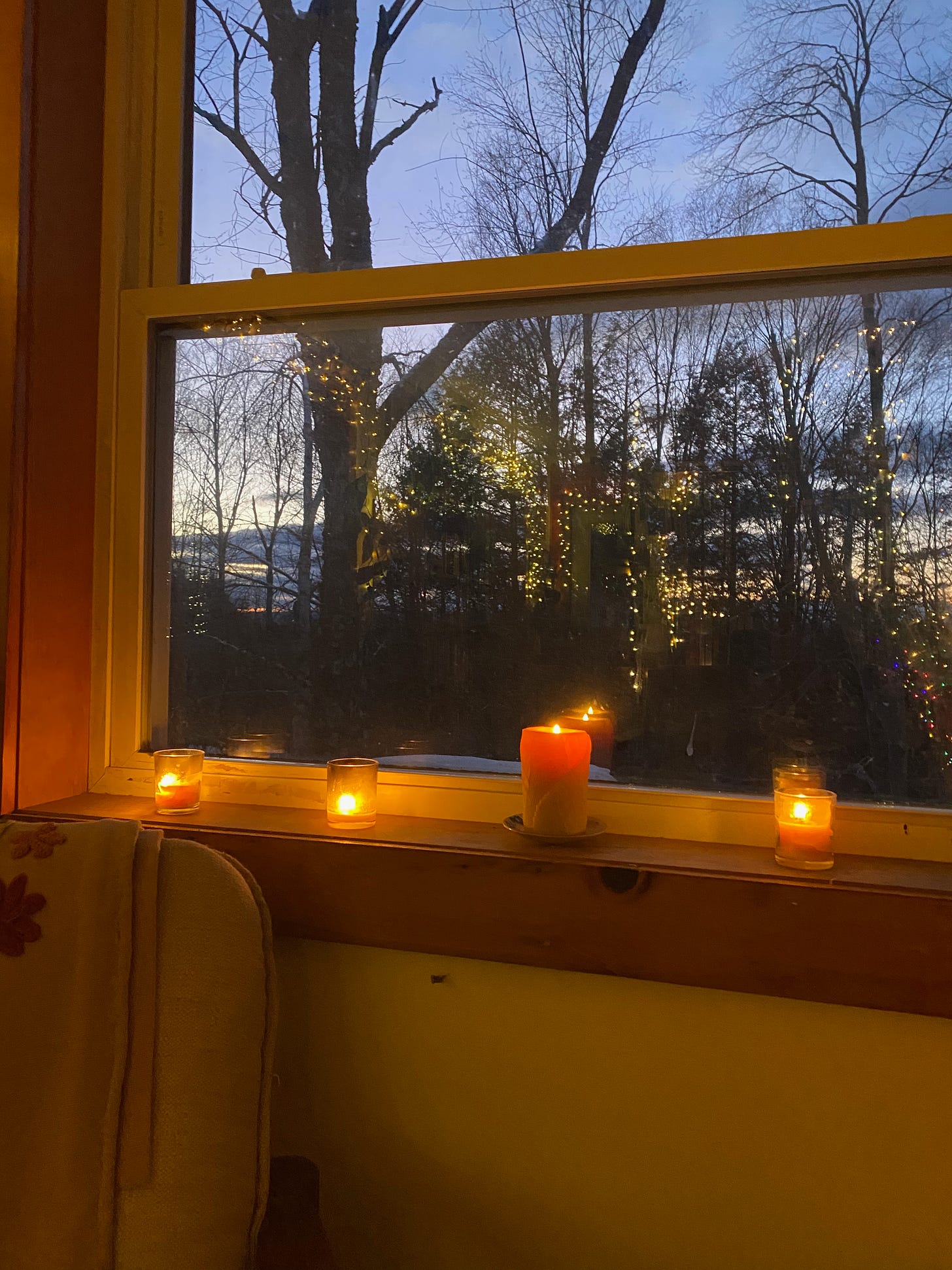 A row of lit candles on a windowsill at dusk; the twinkly lights from around my house are reflected in the window.