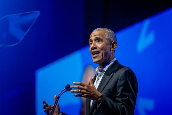 Former President Obama, standing at a microphone in front of a blue background, gestures with his hands as he speaks.