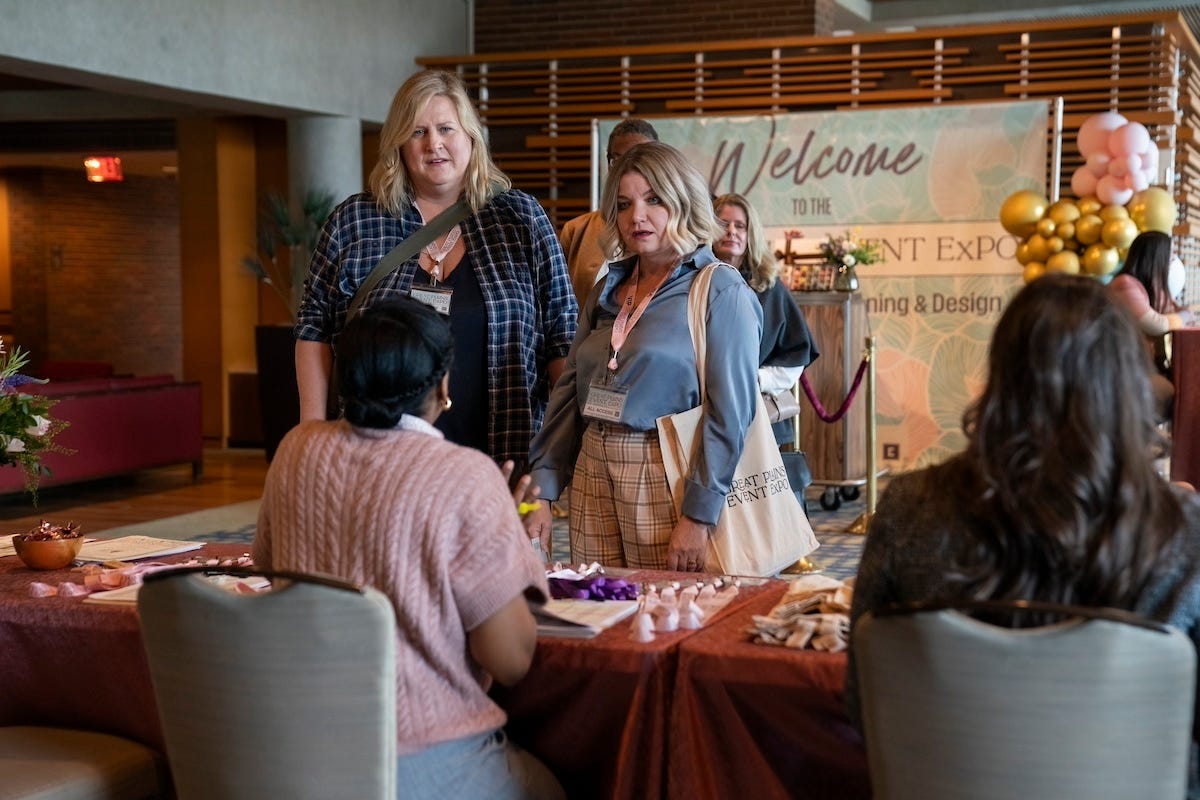 Bridget Everett and Mary Catherine Garrison are standing in front of a registration table on the set of Somebody Somewhere