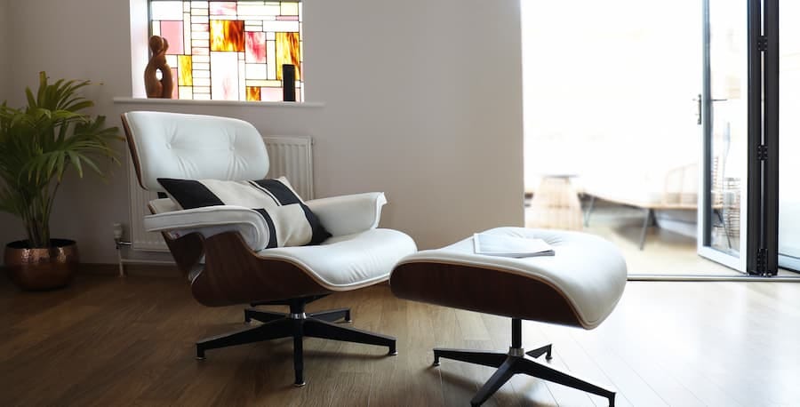 The living room has a white leather armchair with a matching ottoman on a wooden floor. There is a large stained glass window on the left wall with a plant in front of it. There are large bi-fold doors positioned to the right leading into a conservatory.