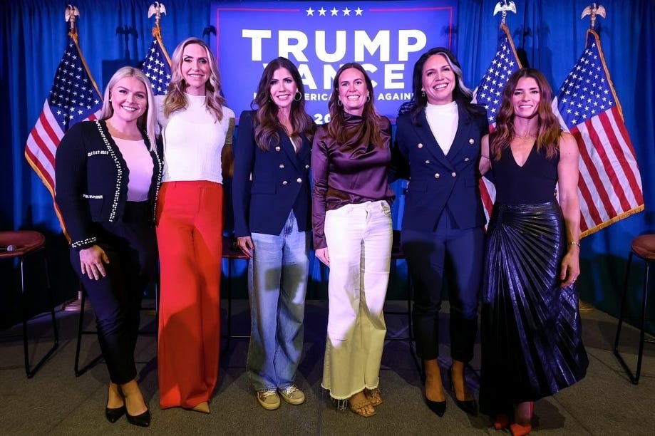 Group of six women standing together at a Trump campaign event with American flags in the background.