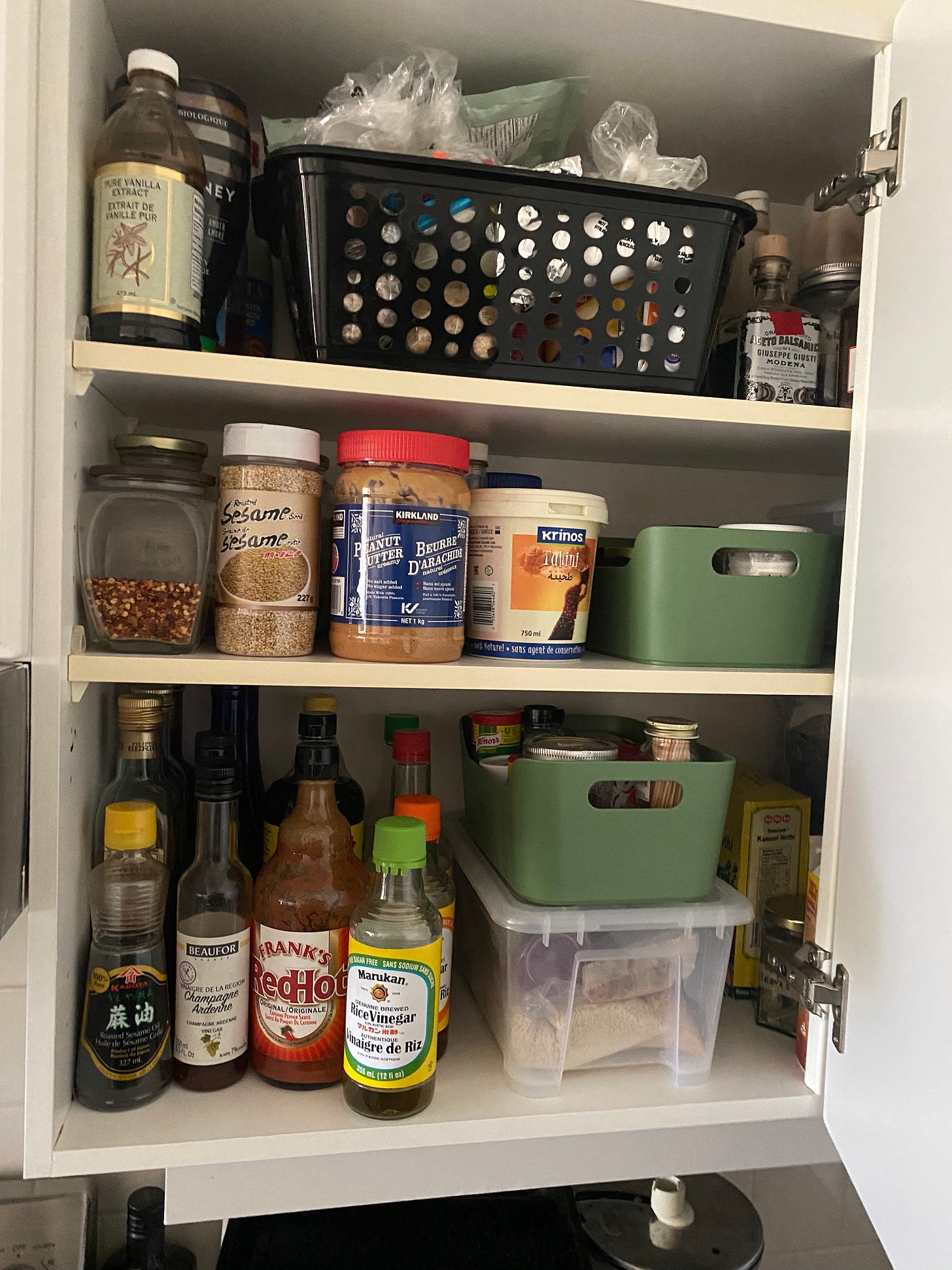 An open cupboard filled with pantry items: oils and vinegars, baking supplies, nut butters, etc. Things are neatly lined up in rows interspersed with clear containers, baskets, and small but deep mint-green trays with handles.