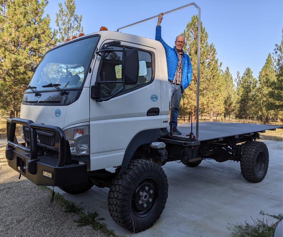 Man standing on the back of a white work truck, holding a single metal rib that represents the outline of the camper yet to be built