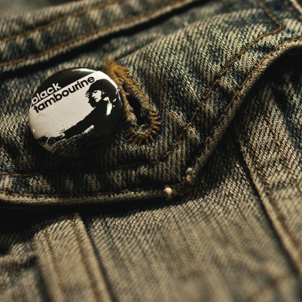 Black Tambourine album cover, with a macro mode photo of a faded denim jacket pocket that has a small black tambourine button pinned to it