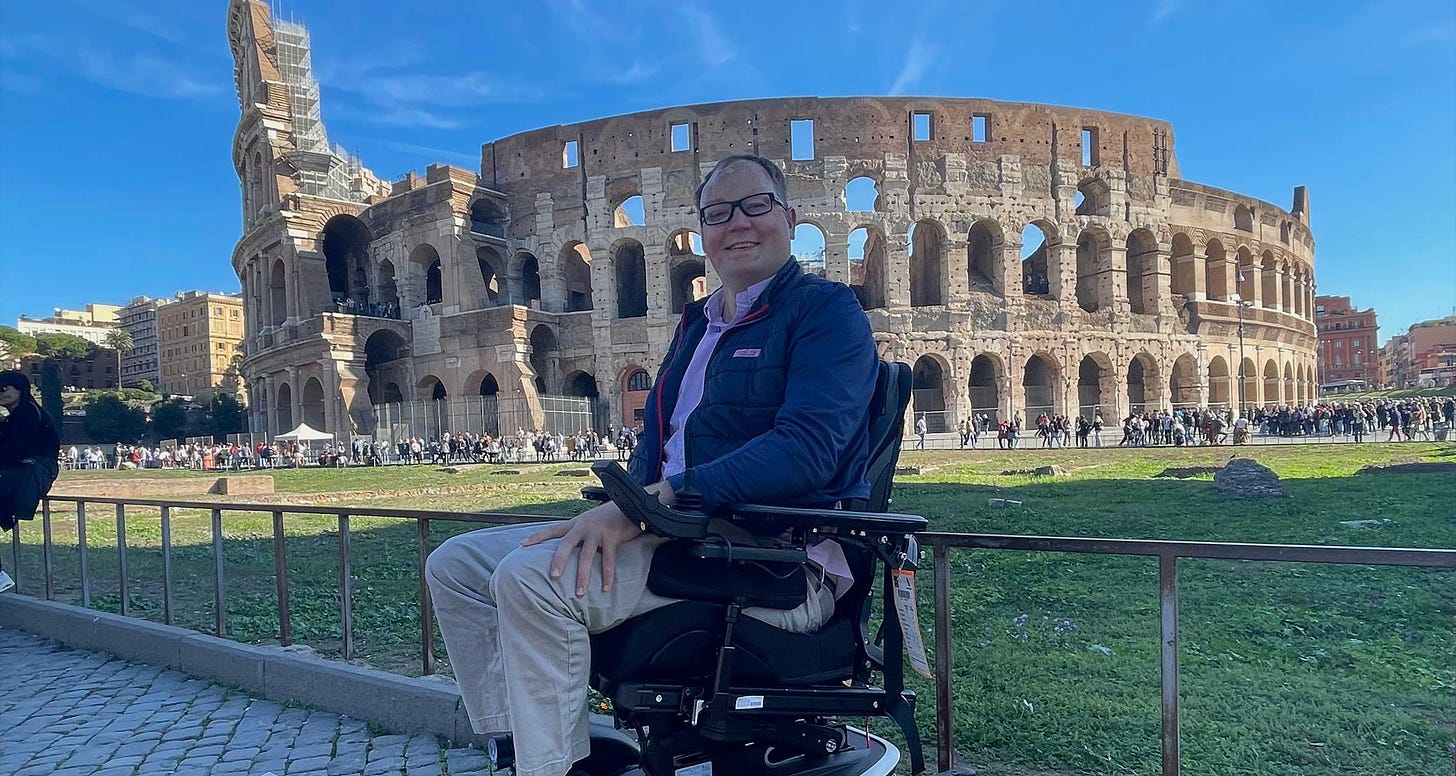 John seated in his wheelchair in front of the Roman Colosseum.