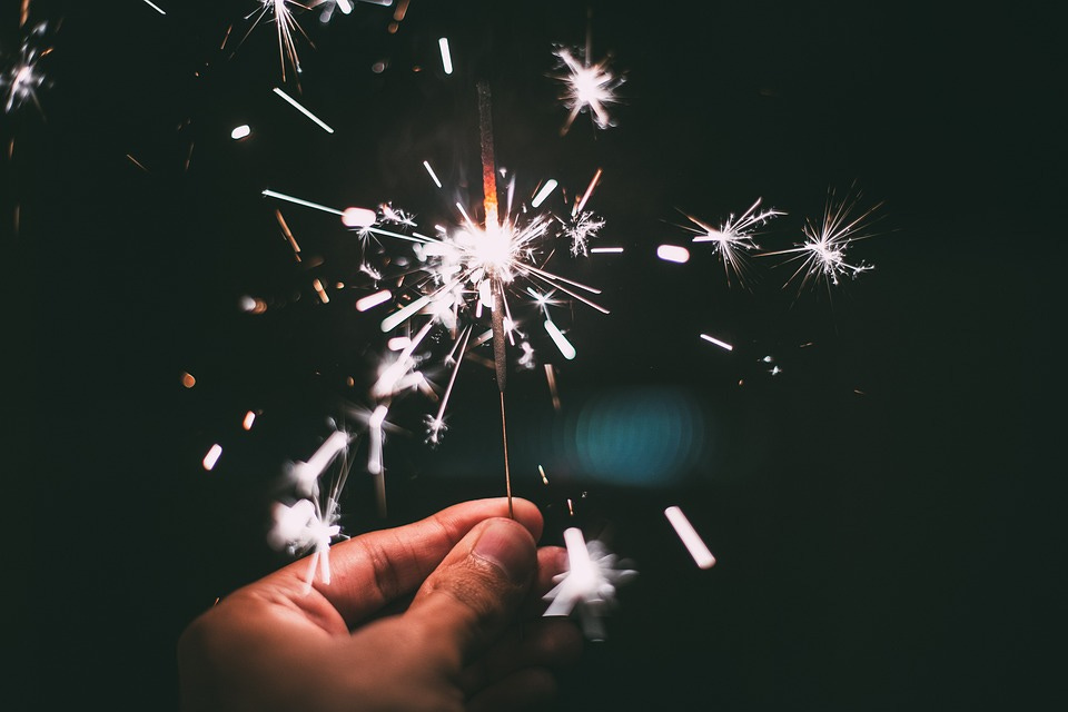 Foto de uma mão segurando uma vela do tipo estrelinha, queimada pela metade e soltando faíscas claras que, em contraste com o fundo escuro, lembram fogos de artifício.