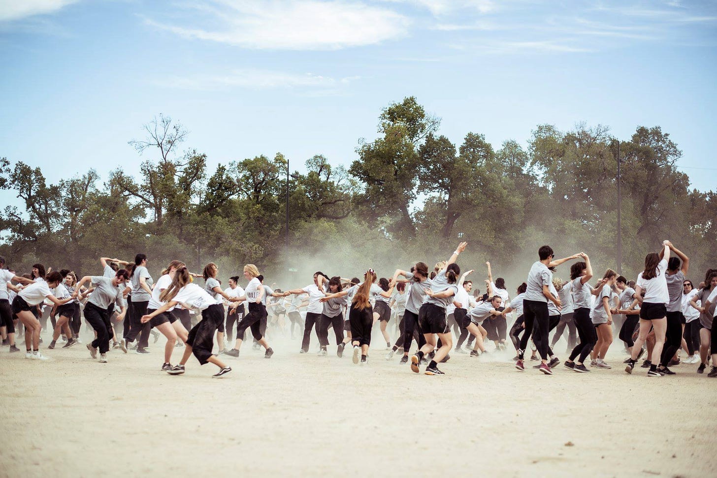 A group of dancers spin on gravel at Birrarung Marr. The dancers wear black pants, white or grey t-shirts, and runners. Dust rises from the gravel and there are gum trees in the background.