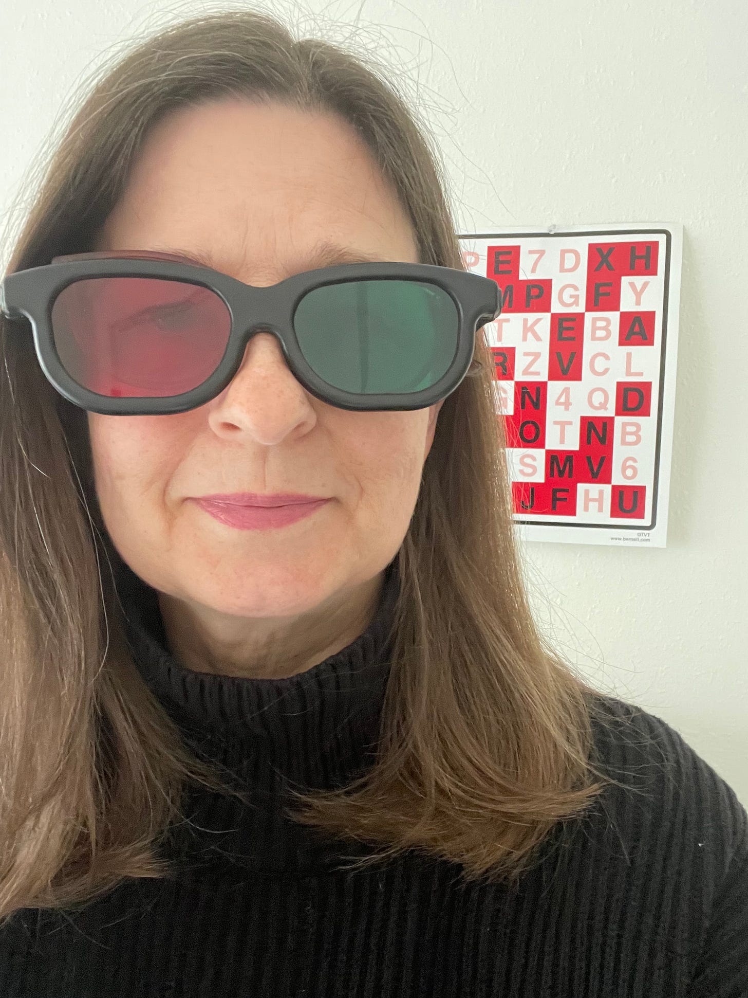 Woman standing in front of eye chart, wearing glasses. One lens of glasses is red, and the other is green.