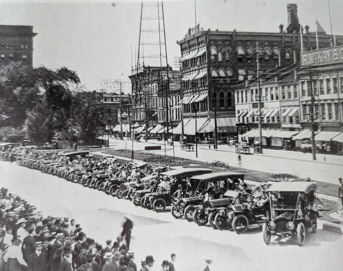 A line of cars from the early 1900s in front of Hamilton's Gore Park with lines of spectators looking at the vehicles.