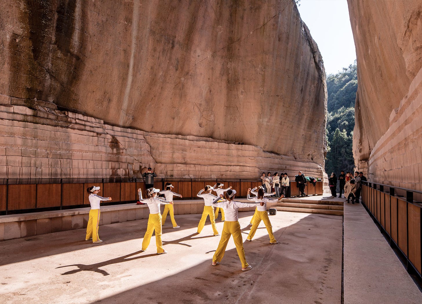 Dancers in yellow pants and white shirts perform on a recessed rectangular "stage" set within a rock quarry; beyond them, visitors watch the performance and a narrow opening reveals trees and a hill in the background Dancers in yellow pants and white shirts perform on a recessed rectangular "stage" set within a rock quarry; beyond them, visitors watch the performance and a narrow opening reveals trees and a hill in the background